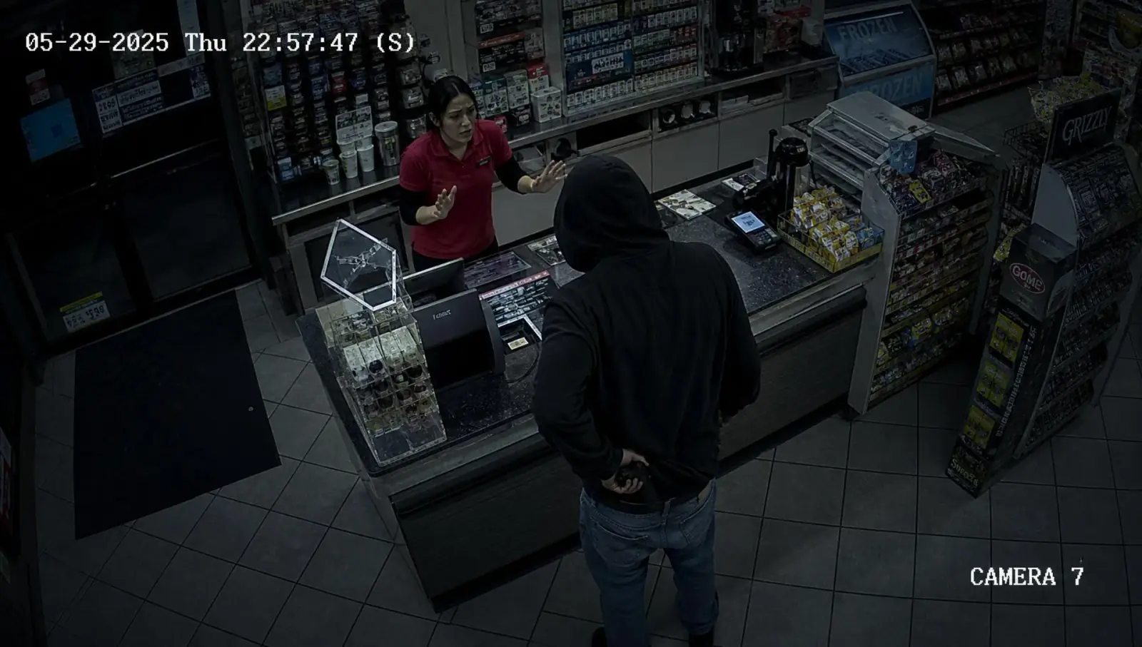 Security camera footage of a hooded suspect drawing a concealed weapon in front of a convenience store counter while an employee stands behind it